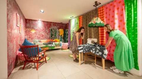 Phil Wilkinson A room setting in the exhibition shows a colourful patchwork sofa with lots of different patterns, in a room with red Oriental-style floral wallpaper and a wall of curtain panels of very different bold patterns. Two women are lifting a sewing table into place.