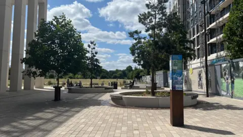 Brafa Square with benches surrounding trees and a column with information plaques around it. Shoreditch Park is behind the square.