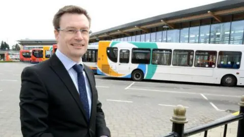 Stagecoach Stagecoach East Midlands Managing Director Matt Cranwell standing to the left of the photo. He is middle-aged wearing a black blazer, light blue shirt and navy tie with white spots. He is smiling, has dark hair and is wearing silver thin framed glasses. He is in a coach park, with a row of yellow, white and turquoise single decker buses behind him.