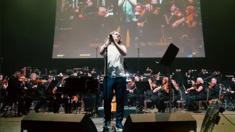 Zoe's Place Jamie Webster is wearing jeans and a white T-shirt as he sings on stage, with the orchestra behind him. An image of him and the orchestra can be seen on a large screen behind them.