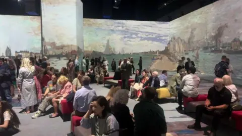 Jermaine Foster/BBC Many people sitting down to view projected images of Monet's Impressionist paintings, depicting ships in a harbour.