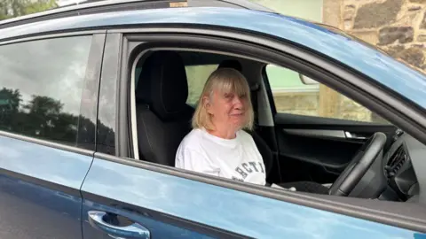 Sheila McPheely sitting in the driver's seat of a blue car. She is smiling at the camera. She has blonde hair and is wearing a white T-shirt with grey writing on it.