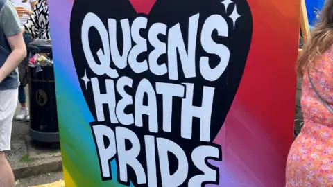 BBC A large colourful piece of artwork spells Queens Heath Pride in white writing on a black heart, with a rainbow background