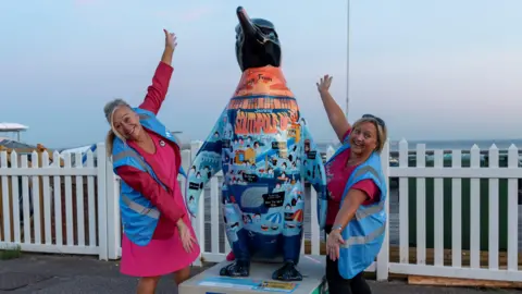 Main Media Two people are standing either sides of a penguin sculpture which is in the middle. Both women have blonde hair and are wearing a a blue hi-vis jacket standing in front of a white fence.