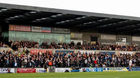 SNS A general view of a stand at Somerset Park. Hundreds of fans are tightly packed into a stand covered in advertising hoardings. There are some players and match officials in the foreground on the pitch. 