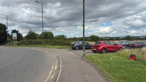 A layby near a roundabout at Edinburgh Airport. There are four cars. The car closest to the camera is red. The cars further away are dark coloured, either black or grey.