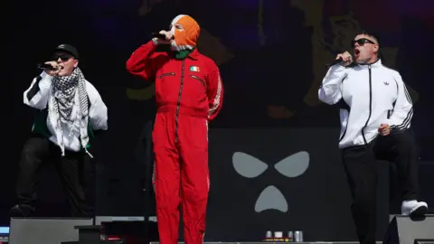 PA Media The band Kneecap perform onstage - all three members of the band are holding microphones. One of the group, standing centre, is wearing all red with a small Irish flag printed on it and a tricolour balaclava. The other two band members are wearing white jackets and sunglasses.