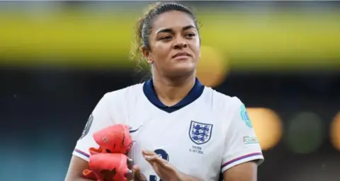Getty Images Jess Carter playing for England in a white and navy blue shirt. She looking into the distance while clapping with her pink football boots tucked under her left arm.