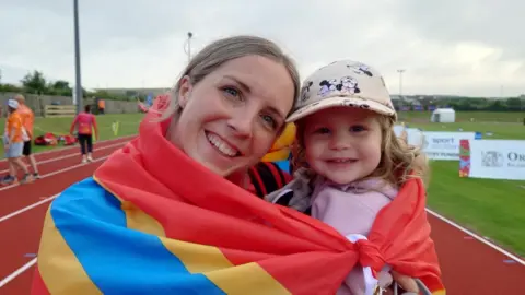 David Griffiths A woman holding a small girl, wrapped with a red, blue and yellow Orkney flag