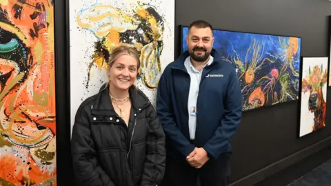 Guernsey Ports Tiffany Anna and David Buddery stand in front of Tiffany' artwork in the terminal.