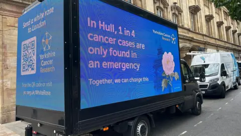 BBC News A blue van displaying digital billboard for Yorkshire Cancer Research is parked outside a grand, stone-built city hall. Messages read: "Send a white rose to London", and "In Hull, one in four cancer cases are only found in an emergency - together we can change this".
