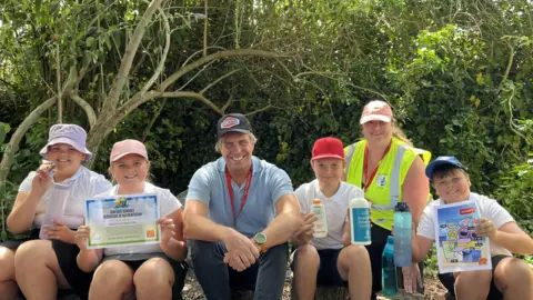 BBC Charlotte, 10, Hattie, 9, Tony Tostevin (MUG), Ollie, 10, Juliet Bell (Vale Primary School Sun Safety Ambassador teacher) and Lewis, 9, sat in a shaded woodland area at the school. They are all smiling, wearing sun hats and holding an array of items like suncream, the sun-safe accreditation certificate and waterbotles. 