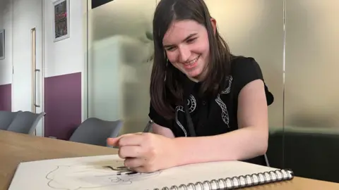 BBC Lila in an office, smiling with her head down focused on drawing a cartoon character. She is smiling and has long black hair and is wearing a dark top.