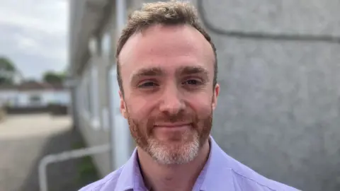 Younger man with beard and moustache and flouncy hair smiling at the camera