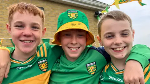 BBC Three young boys in Donegal GAA gear. The boys on the left and right are wearing yellow and green county jerseys while the boy in the middle, who has his arms around the other two, is wearing a green Donegal jumper and green and yellow bucket hat.