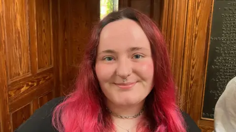Ciara Kennedy has long dyed pink hair and blue eyes. She is standing in front of a wooden door smiling. She is wearing a silver choker necklace and finer silver chain below.
