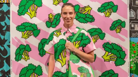 Phil Wilkinson Designer Ida Pettersson Preutz smiles while wearing her broccoli pattern in a shirt against a backdrop of the same fabric and holding a tote bag in the same bright pink and white stripes with large green florets