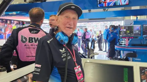 Lucy Wallduck An older man wearing a black cap, blue headphones, and a black shirt stands in a motorsport pit area. Behind him, team members in pink and black uniforms work near monitors, with spectators visible through the open garage.