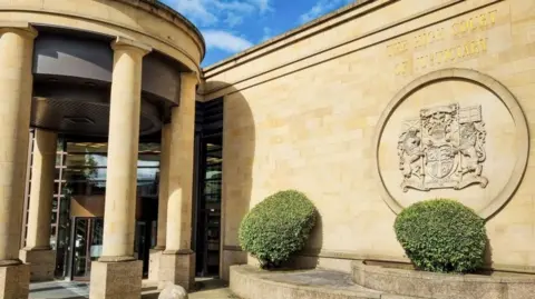 Getty Images Exterior photograph of the front and entrance to the High Court in Glasgow. The sand-coloured building features a large stone-carved coat of arms above one of two hedges to the right of the entrance, which is a glass panelled revolving door.