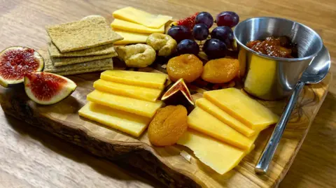 BBC A selection of cheeses, figs, dried apricots, nuts, grapes, chutney and crackers sits on a wooden cheeseboard on a table.