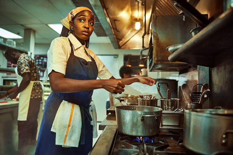 A young black woman chef standing at a stove looking watchful and nervous.