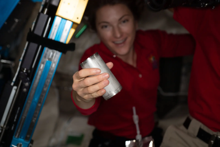 An astronaut in a red shirt holds a small metal cylinder.