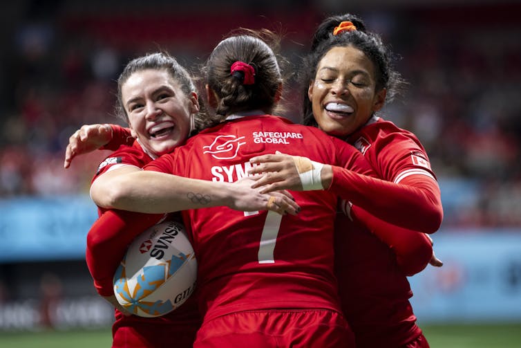 Three women in red rugby uniforms embrace on a rugby pitch