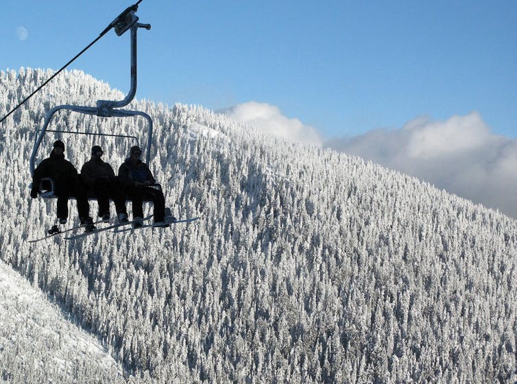 Three people on a ski lift silhouetted against a hilliside of snow-covered evergreen trees and a blue sky