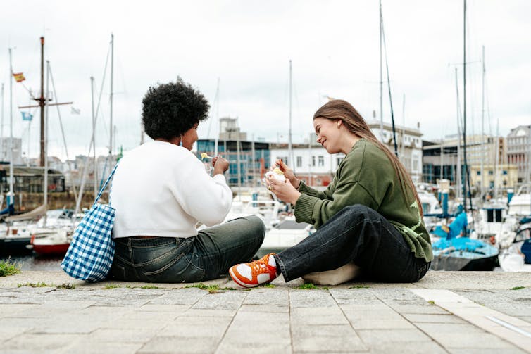 Two young women sitting on a dock near a marina on a cloudy day