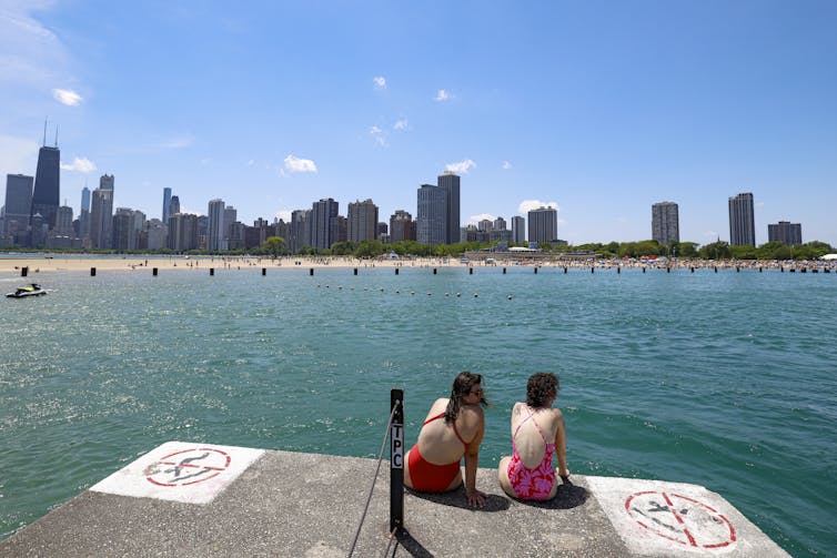 People sit on a concrete pier sticking out over an area of water, with tall buildings in the background.