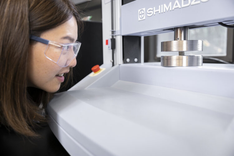 A researcher in safety glasses observes a small cement cube sandwiched between two metal discs in an industrial-looking machine.