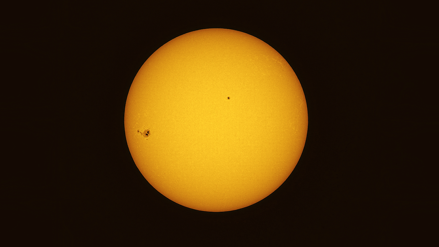The sun's yellow disk is shown against a black background. A collection of sunspots can be seen to the left of the otherwise placid face of our star, with another close to its center.