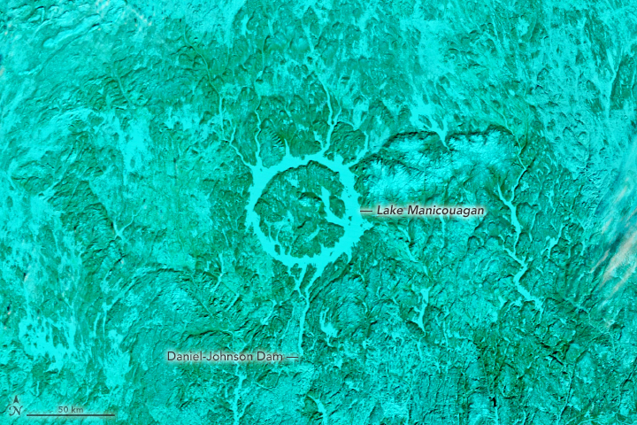 A false-color version of the image shows the snow-covered vegetation as greenish-blue. Snow and ice appear blue, including the ice that covers the ring-shaped lake centered in the image.