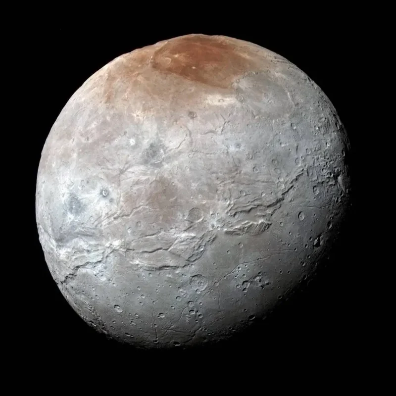 A detailed image of Charon, Pluto’s largest moon, showing a gray, cratered surface with a reddish-brown area at the north pole and visible ridges and valleys, set against a black background.