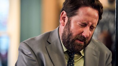 Jim Howick in character as Paul wincing. He is wearing a grey suit and tie with a lemon yellow shirt.