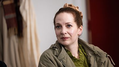 Katherine Parkinson in character as Rachel wearing a khaki green coat with her hair in a bun.