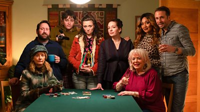 A group of 8 people sat and stood around a poker table covered with chips. A few of them are holding drinks, one is holding a video camera, and another is holding a small plastic winners trophy cup.