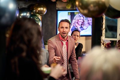 A man in a pink suit and tie is pointing at somebody out of shot. There are balloons around him.