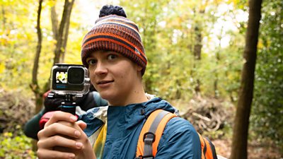 A young adult wearing a woolly bobble hat in a forest. He is pointing a video camera at the camera.