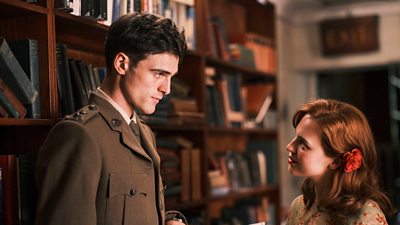 A young man in military uniform talking to a young woman with a flower in her hair. They are in a room which has a large bookcase filled with old books.
