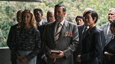A group of ten people stood in smart dark clothing. The central character appears to be holing a religious book and is wearing his military medals on his suit.