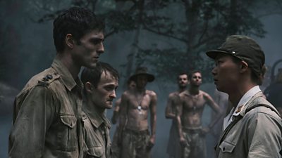 Men stood in a dark setting looking solemn. Some are in military uniform whilst others are not wearing clothing on their upper half