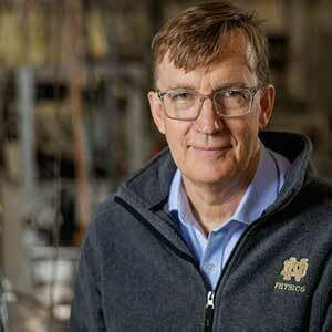 Graham Peaslee, a white man, is pictured wearing a gray quarter zip pullover featuring the ND Physics logo in gold on the left side of the chest. He has short, light brown hair and glasses. His lab is pictured in the background, out of focus.