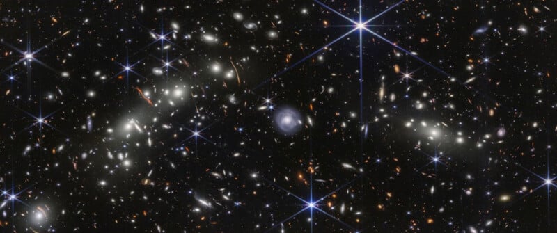 A deep space image showing countless distant galaxies of various shapes and sizes, scattered against a dark background with several bright stars featuring diffraction spikes.