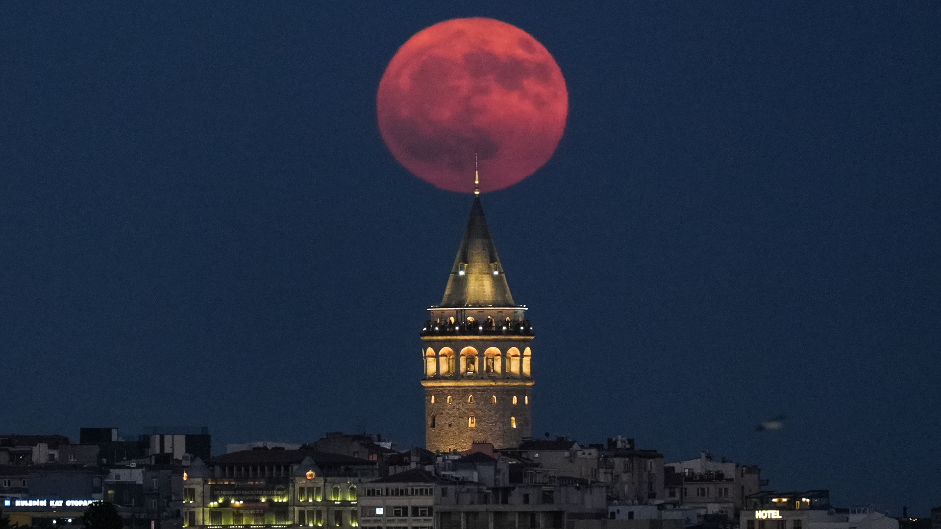 A reddish full moon is pictured above a castle-like tower lit from within with golden light during night time.