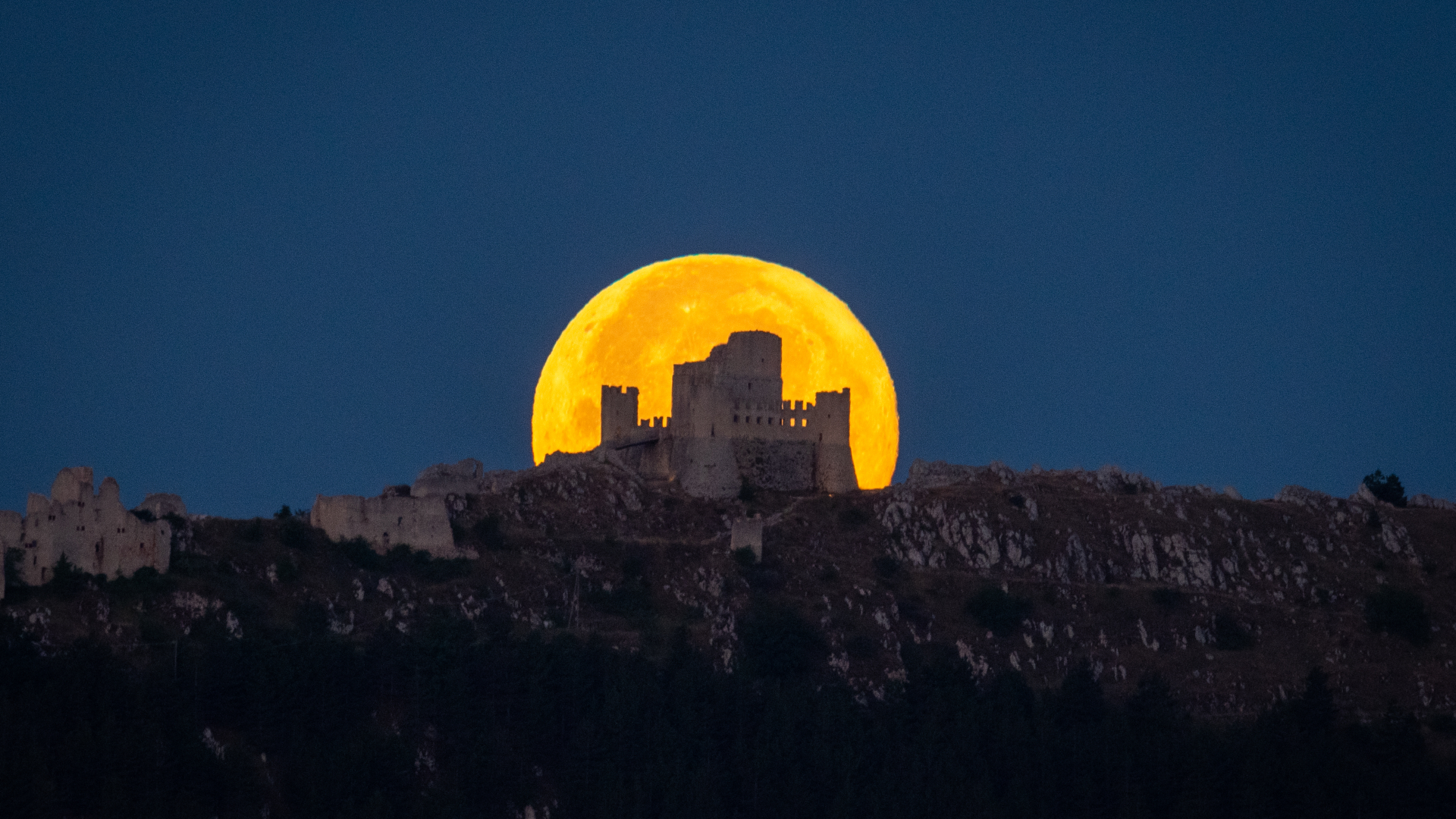 An orange full moon can be seen setting behind the ruins of a castle on a rocky hillside.