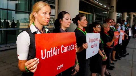 Carlos Osorio / Reuters Air Canada flight attendants picket at Toronto Pearson airport holding signs that read "unfAir Canada" and "unpaid work won't fly". They are standing in a line and wearing their uniforms. 


