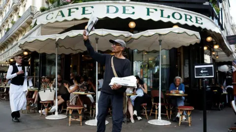Reuters Ali Akbar, wearing a grey flat cap and a black shirt, stands with a paper held high in his right hand in front of the Cafe De Flore in Paris