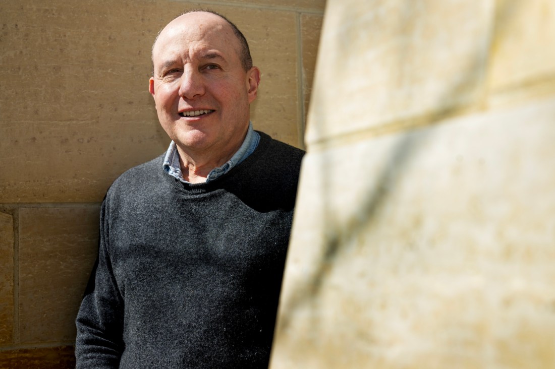 Peter Mancusi, associate teaching professor of journalism at Northeastern University, stands smiling in sunlight against a stone wall, wearing a dark gray sweater and blue collared shirt.