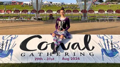 Eilidh Gammons The image shows a young girl, wearing a purple and white highland dance costume, sitting on a banner that reads "Cowal Gathering 29th-31st Aug 2024". She is holding a trophy.